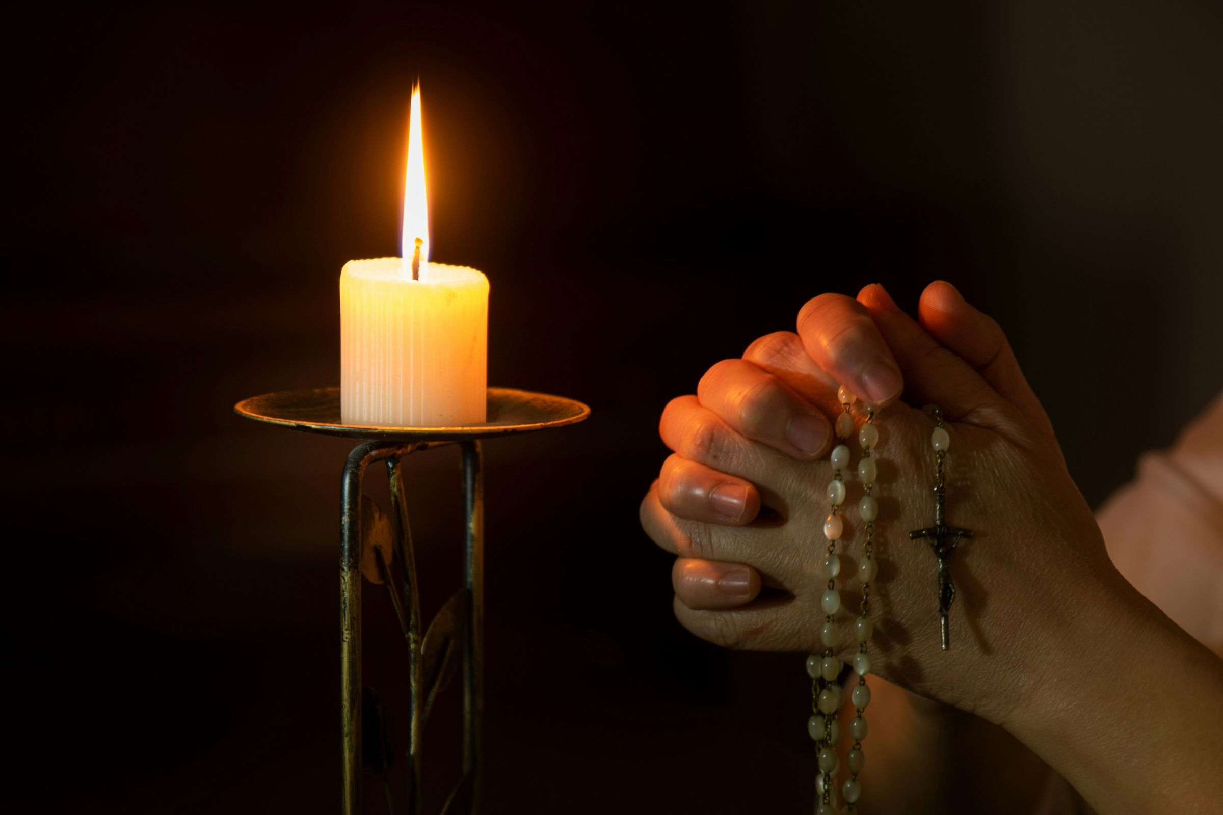 Close-up of hands holding a Rosary with a candlelit background