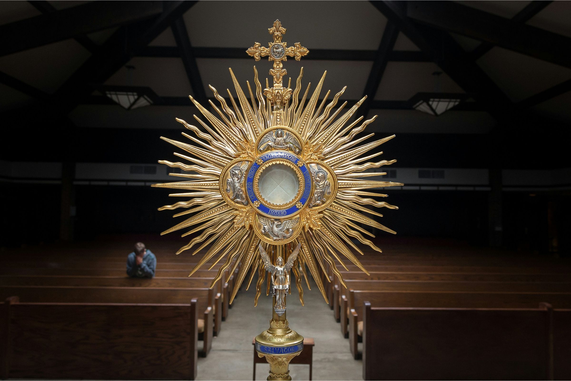 Close-up of the Eucharist in a golden monstrance on an altar with a young man praying in the background